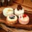 Four assorted mini cheesecakes from The Watering Can Pastry Market, displayed on a wooden board with colourful toppings including fresh berries and cream.
