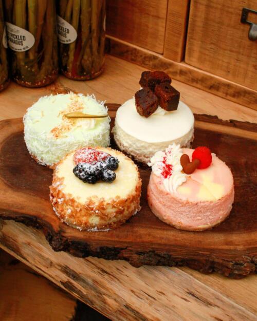 Four assorted mini cheesecakes from The Watering Can Pastry Market, displayed on a wooden board with colourful toppings including fresh berries and cream.