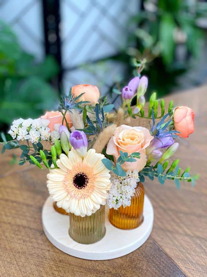 The Watering Can | A set of 4 orange and beige glass vases no a round wooden tray filled with blush peach and purple flowers.