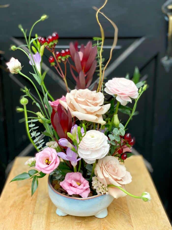 The Watering Can | An elegant european arrangement of blush, lavender, and burgundy blooms in a shallow light blue dish.