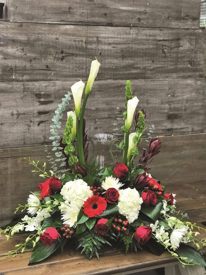 The Watering Can | White, red, and burgundy flowers and greens arranged all around an urn.