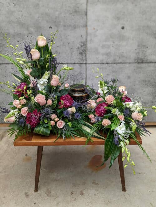 The Watering Can | A spectacular blush, plum and blue creamtion design. Soraing above the urn on the right, the arrangement flows around the urn, forwads and down again on the left.