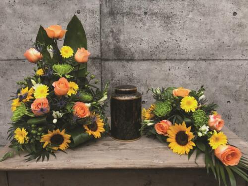 The Watering Can | A peach and yellow cremation display featuring sunflowers, roses, mums, and gerbera daisies.