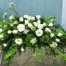The Watering Can | An all white casket spray made with bells of Ireland, gerbera daisies, mums. roses, ranunculus, trachelium, queen anne’s lace, waxflower, and curly willow in a lush bed of greens.