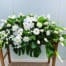 The Watering Can | An all white casket spray made with calla lilies, roses, dendrobium orchids, mums, star of Bethlehem, ranunculus, orchids, and trachelium in a bed of tropical greens.