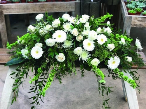 The Watering Can | An all white casket spray made with bells of ireland, roses, mums, gerbera daisies, trachelium, ranunculus, and waxflower in a bed of lush greenery.