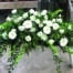 The Watering Can | An all white casket spray made with bells of ireland, roses, mums, gerbera daisies, trachelium, ranunculus, and waxflower in a bed of lush greenery.