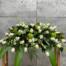 The Watering Can | A casket spray made with white roses, white dendrobium orchids, green hydrangea, and burgundy hypericum in a bed of lush greens.