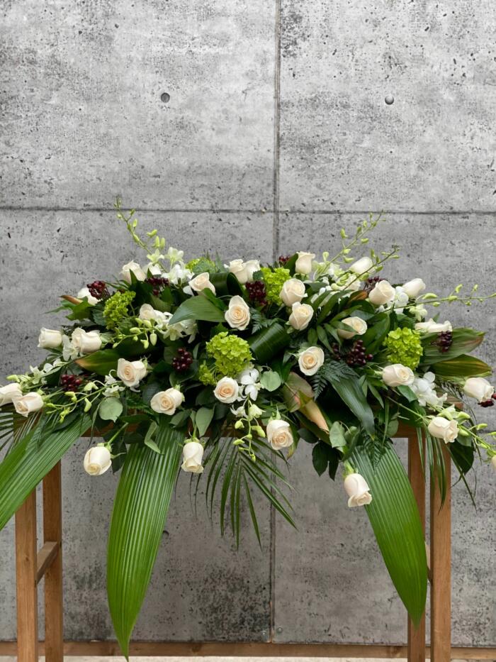 The Watering Can | A casket spray made with white roses, white dendrobium orchids, green hydrangea, and burgundy hypericum in a bed of lush greens.