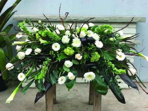 The Watering Can | A green and white casket spray made with green hydrangea, white calla lilies, white roses, bells of Ireland, white ranunculus, white gerbera daisies, green hypericum, and white mums in a lush bed of greens.