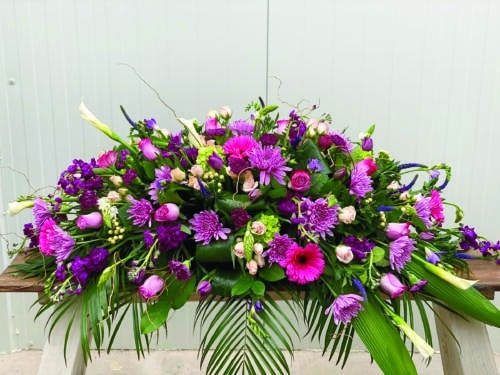 The Watering Can | A purple casket spray made with white calla lilies, purple roses, lavender mums, green hydrangea, dark purple stock, soft pink spray roses, stars of Bethlehem, purple freesia, purple veronica, hot pink gerberas, purple tulips, and white hypericum on a bed of greens.