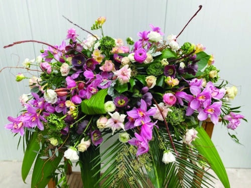 The Watering Can | A large and luxurious purple casket spray made with pink cymbidium orchid, pink roses, soft pink freesia, dark purple helleborus, ranunculus, butterfly ranunculus, and white anemone on a lush bed of tropical greenery.