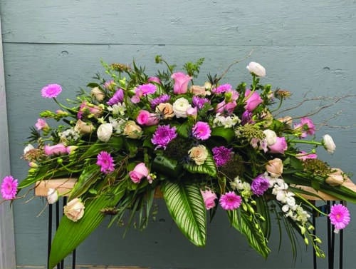 The Watering Can | A large pink and purple casket spray made with soft pink roses, lavender mums, thistle, chocolate queen anne’s lace, purple trachelium, soft pink freesia, white ranunculus, soft pink gerberas, and white dendrobium orchids in a bed of tropical greens.