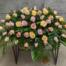 The Watering Can | A casket spray made with soft pink, soft yellow, and peach roses supported by waxflower and a variety and greens.