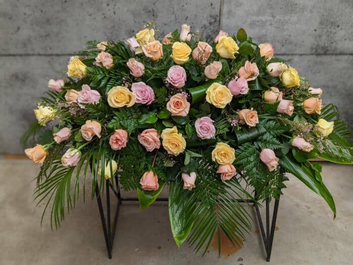 The Watering Can | A casket spray made with soft pink, soft yellow, and peach roses supported by waxflower and a variety and greens.