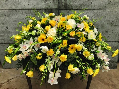 The Watering Can | A yellow casket spray made with white cymbidium orchid, bright yellow roses, white peonies, bright yellow gerberas, white dendrobium orchids, stars of Bethlehem, yellow freesia, green hypericum, and white trachelium on a bed of greens.