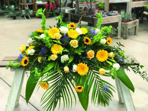 The Watering Can | A casket spray made with purple agapanthus, bells of Ireland, sunflowers, yellow gerberas, soft yellow roses, and white roses on a bed of greens.
