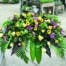 The Watering Can | A colourful caskey spray in purples and yellows made with soft yellow roses, bells of Ireland, pink lisianthus, lime green hydrangea, dark purple calla lilies, lavender mums, bright yellow gerberas, purple freesia, pink tulips, star of Bethlehem, and peach ranunculus on a bed of greens.