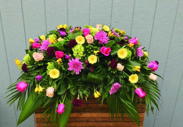 The Watering Can | A pink and yellow casket spray made with hot pink roses, soft pink roses, lavender mums, lime green hydrangea, dark purple moon carnations, dark pink calla lilies, pink tulips, yellow freesia, green hypericum, and soft yellow gerbera on a lush bed of greens.