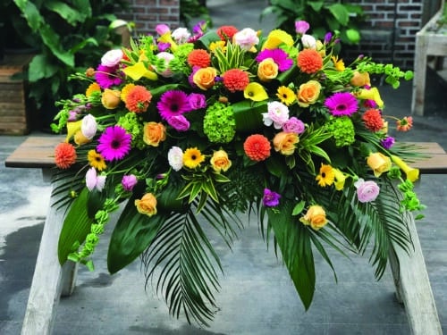 The Watering Can | A bright and colourful casket spray made with butterscotch roses, green hydrangea, yellow calla lilies, hot pink freesia, blush ranunculus, pink lisianthus, yellow gerberas, hot pink gerberas, orange ball dahlias, and bells of ireland on a bed of greens.
