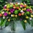 The Watering Can | A bright and colourful casket spray made with butterscotch roses, green hydrangea, yellow calla lilies, hot pink freesia, blush ranunculus, pink lisianthus, yellow gerberas, hot pink gerberas, orange ball dahlias, and bells of ireland on a bed of greens.