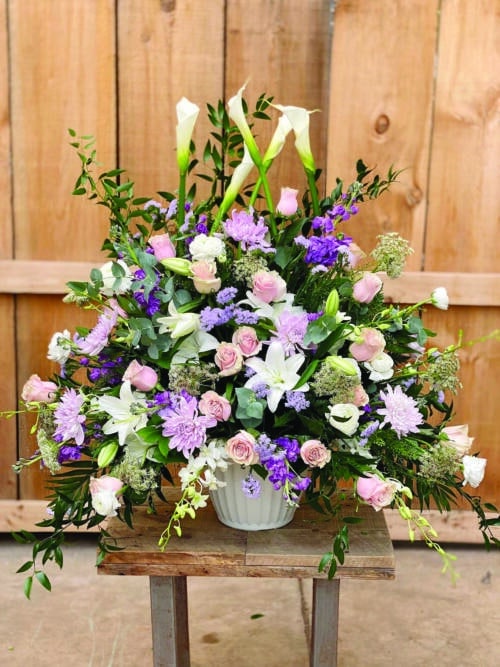 The Watering Can | A stunning large and full side spray made with white calla lilies, soft pink roses, white lilies, lavender mums, blue statice, queen anne’s lace, lavender stock, white ranunculus, and white dendrobium orchids supported by greenery.