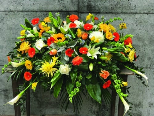 The Watering Can | A warm and bright casket spray made with white calla lilies, yellow spider mums, sunflowers, orange dahlias, yellow/peach dahlias, white Asiatic lilies, orange gerberas, coral reef roses, bells of Ireland, white peonies, yellow freesia, and blue tweedia on a bed of mixed greens.