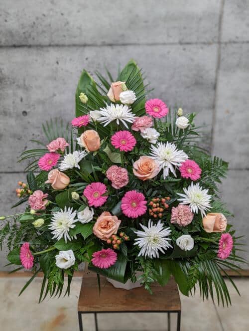 The Watering Can | A large peach, pink, and white side spray made with white mums, shimmer roses, pink gerberas, peach hypericum, dusty pink carnations, and white lisianthus backed by greens.