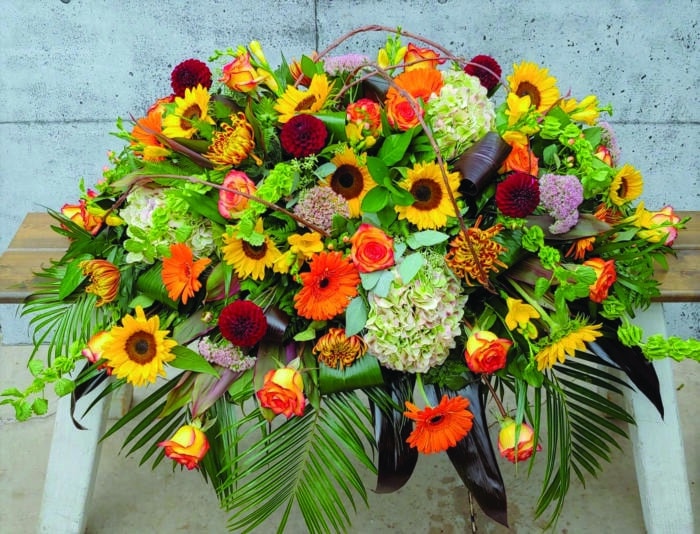 The Watering Can | A round casket spray made with antique hydrangea, orange roses, bells of Ireland, sunflowers, peach hypericum, bronze mums, pink sedum, yellow freesia, burgundy dahlias, orange gerberas, and thin grapevine pieces with a variety of greens.