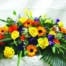 The Watering Can | A colourful casket spray made with yellow calla lilies, yellow roses, orange gerberas, orange dahlia, purple lisianthus, white wax flower, and purple purple freesia on a bed of greens.