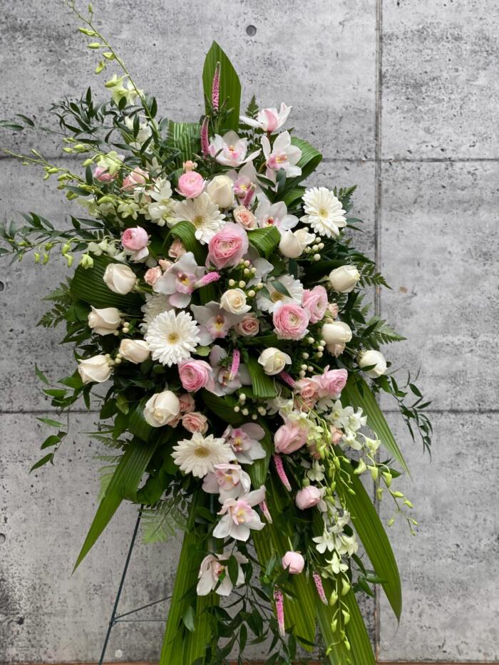 The Watering Can | A blush and white easel spray made with white roses, blush rununculus,soft pink spray roses, white dendrobium orchids, pink veronica, white cymbidium orchids, white gerberas, and white hypericum on a bed of greens.