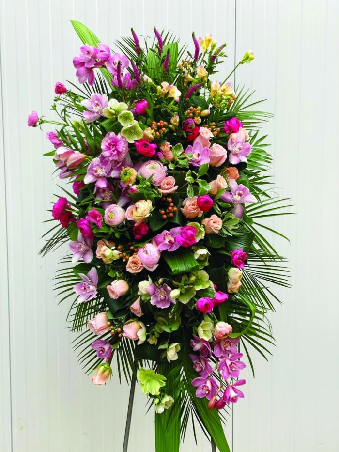 The Watering Can | A large pink easel spray made with roses, helleborus, butterfly rununculus, rununculus, hypericum, cymbidium orchid, freesia, and veronica on a bed of greens.