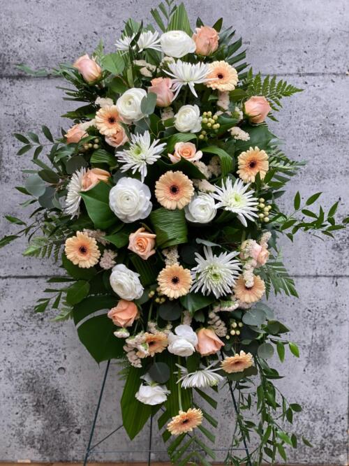 The Watering Can | A peach and white easel spray made with roses, mums, rununculus, hypericum, statice, and gerberas on a lush bed of greens.