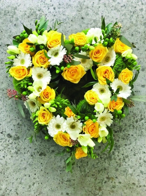 The Watering Can | A small yellow and white open heart floral design with roses, dianthus, gerberas, freesia, and hypericum on a bed of greens.