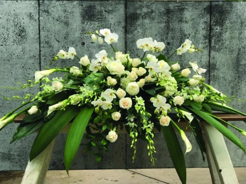 The Watering Can | An all white casket spray made with calla lilies, dendrobium orchids, hydrangea, peonies, roses, and phalaenopsis orchids in a bed of tropical greens.