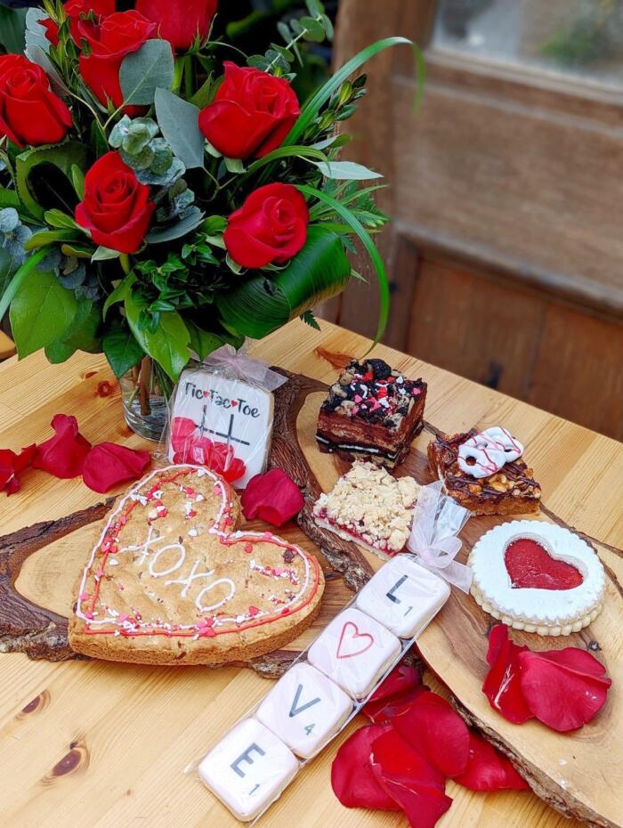The Watering Can | A collection of baked goods with a red rose bouquet in the background.