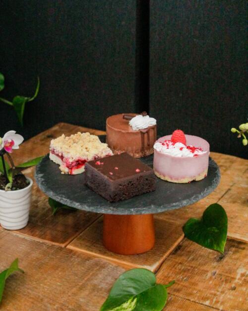 A slate cake stand with a raspberry crumble bar, a chocolate mousse mini cake with whipped topping, a chocolate brownie, and a raspberry mousse cake topped with fresh fruit, with an orchid.
