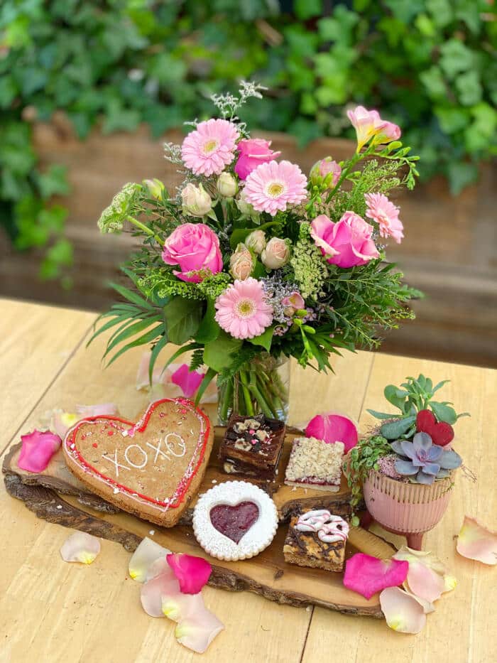 The Watering Can | A pink bouquet and vase, and small succulent planter surrounded by a large heart cookie and other pastries scattered with rose petals.