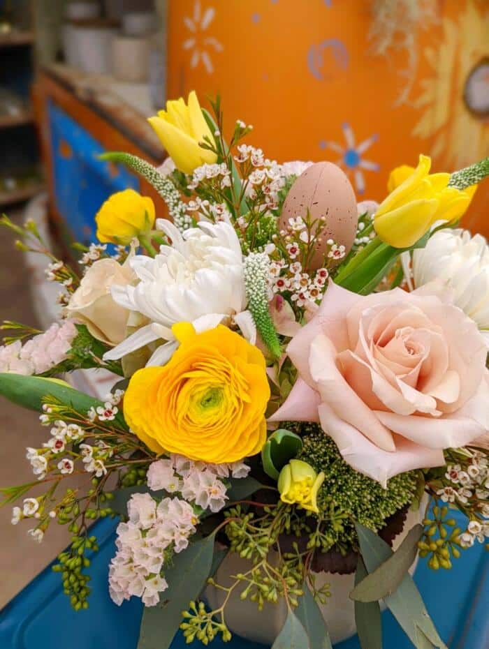 The Watering Can | Close up of a yellow ranunculus, quicksand rose, white mum, and blush statice in a European style floral arrangement.