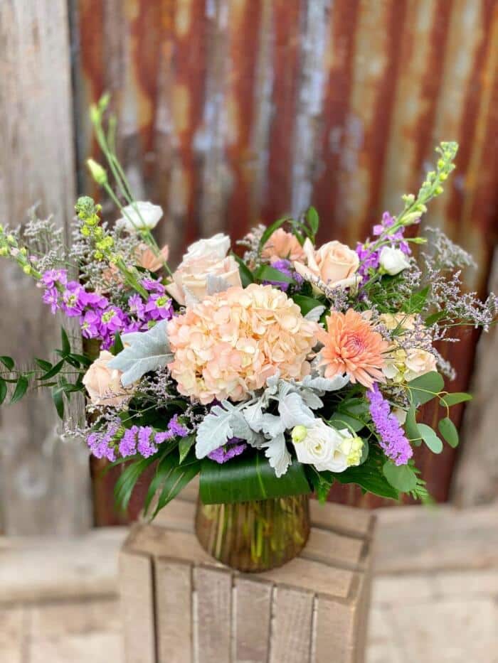 The Watering Can | A peach and purple bouquet in a brown ribbed, glass vase.