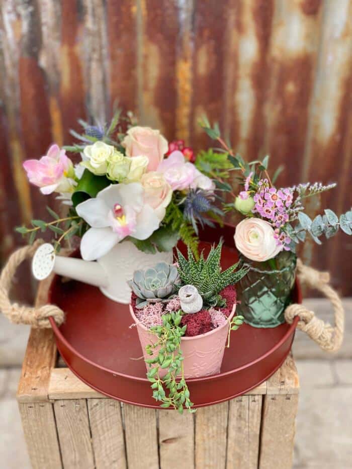 The Watering Can | A burgundy tray with a European arrangement in a white watering can, a planter in a pink tin, and a small glass bud vase with flowers.
