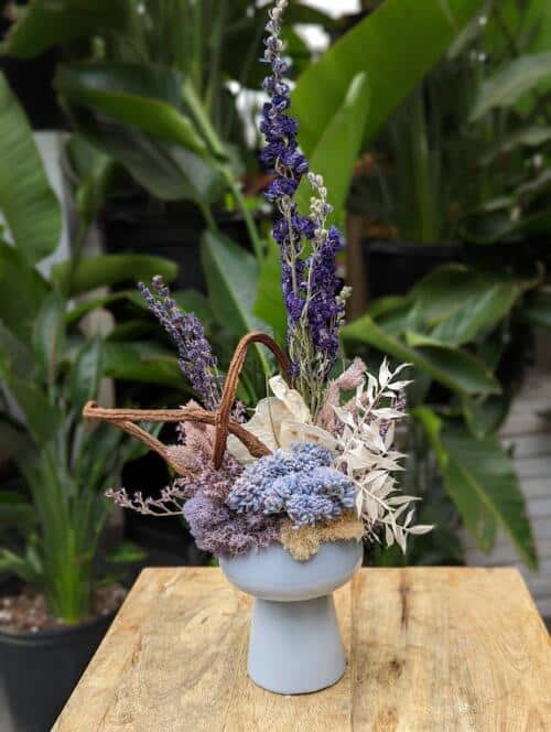 The Watering Can | A funky European style floral arrangement using all dried goods in shades of purple, blue, and natural tones designed in a white ceramic pedestal container.