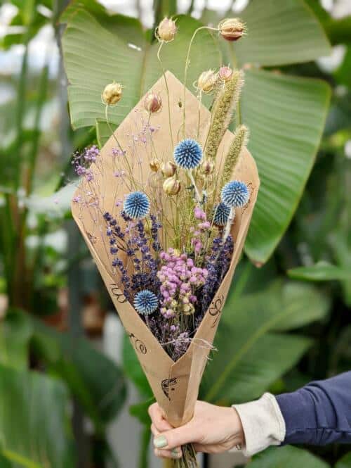 The Watering Can | A bundle of dried flowers in purple, blue, and natural tones wrapped in brown paper.