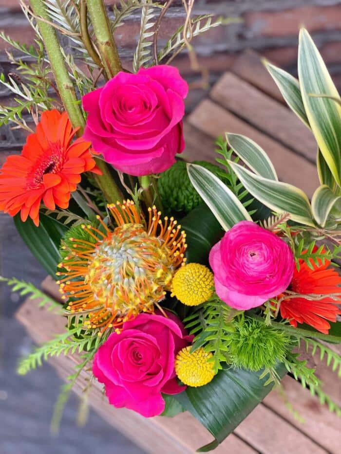 The Watering Can | Hot pink roses, hot pink ranunculus, craspedia, orange gerbera daisies and a pincushion flower in a European style floral arrangement,