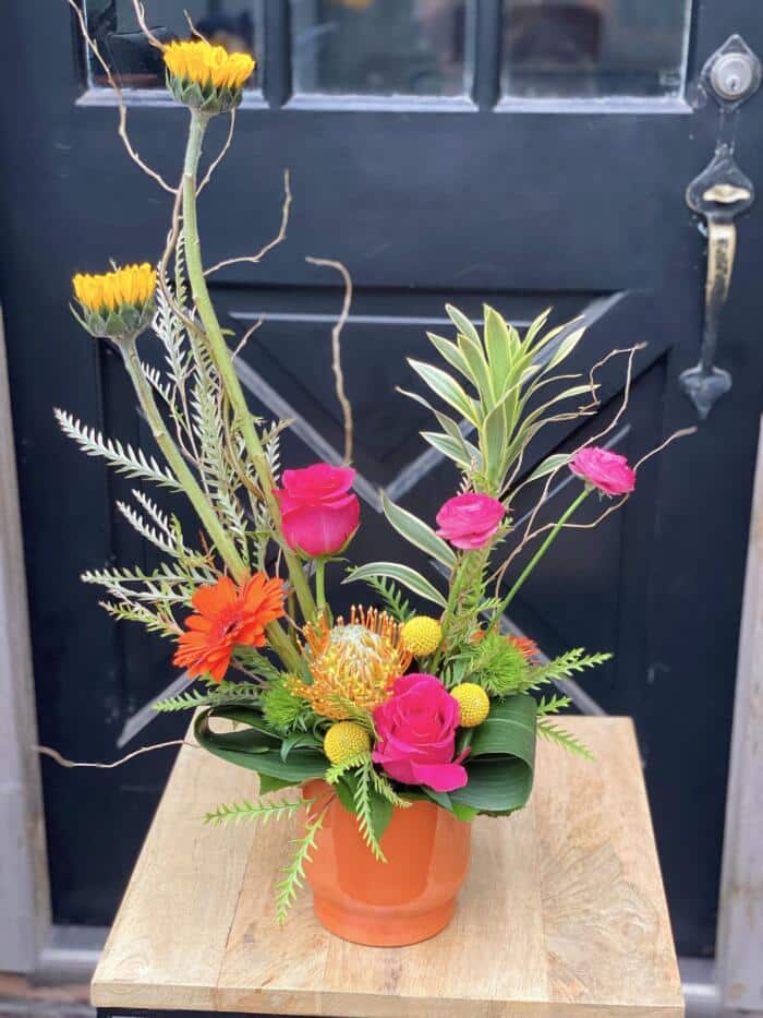 The Watering Can | A bright European style floral arrangement with sunflowers, hot pink ranunculus. hot pink roses, and a pincushion in a bright orange container.