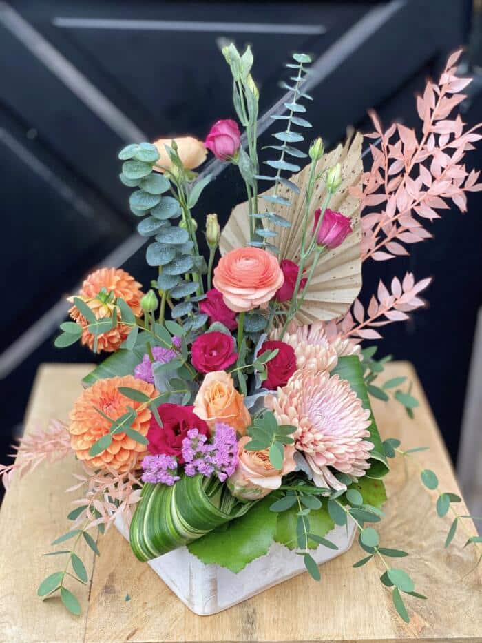 The Watering Can | An profusion of pink, peach, and red blooms amongst textural greenery and dried florals arrange together as an European style arrangement in a white wood container.