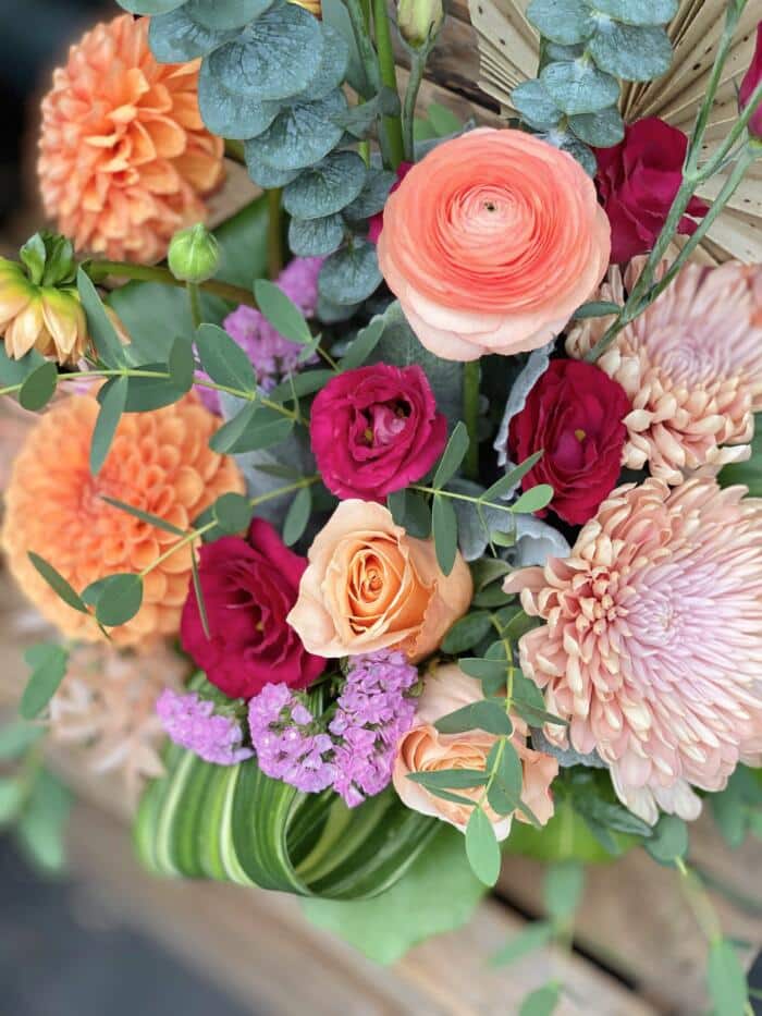 The Watering Can | Orange dahlias, red lisianthus, peach ranunculus, peach mums, and pink statice in a European style floral arrangement.