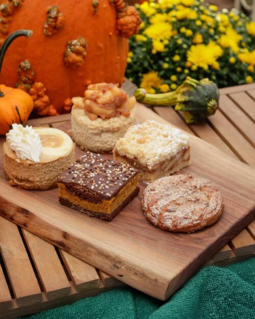 A selection of house-made mini cakes and pastries on a wooden board, styled with an autumn pumpkin and mums.