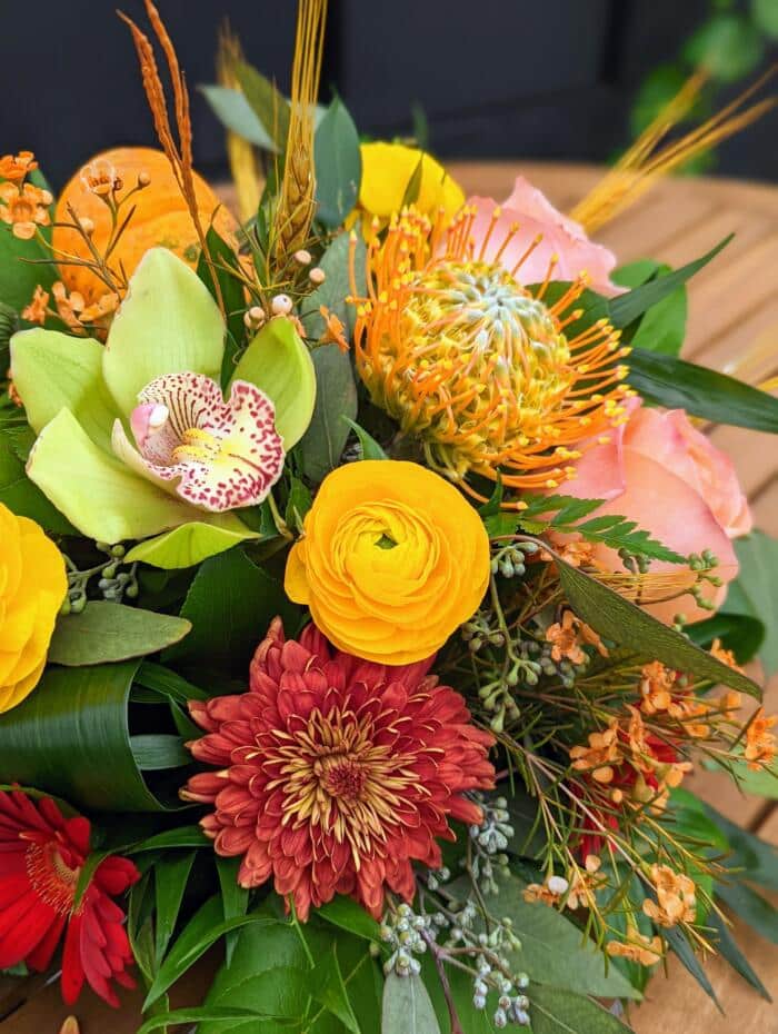 The Watering Can | Close up of green cymbidium bloom, orange mum. yellow ranunculus and pincushion in a table centerpiece arrangement.