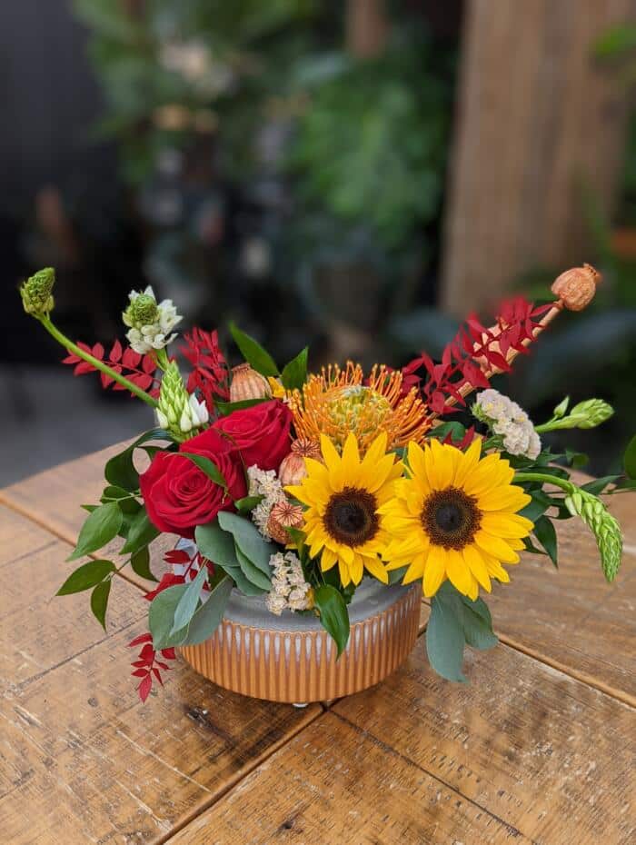 The Watering Can | A low European style floral arrangement featuring sunflowers and bright red roses in a blue and terracotta ceramic container.